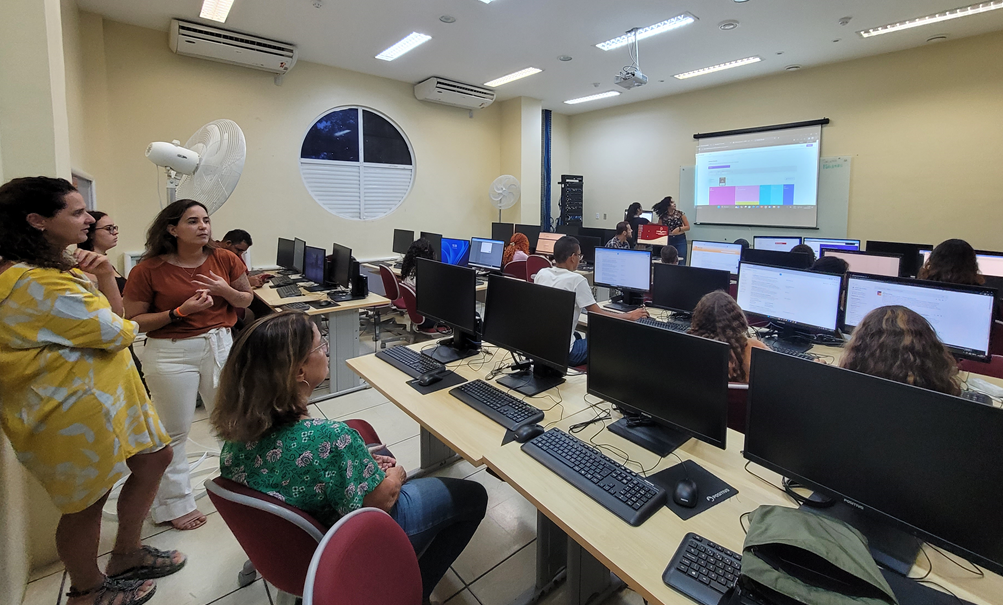 estudantes sentados de frente a computadores e os professores na frente explicando o conteúdo.