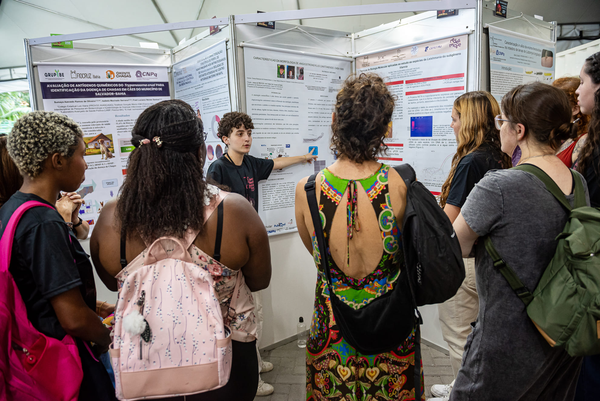 Estudante do ensino médio apresenta seu projeto de pesquisa em um painel de pôster científico para um grupo de pessoas, durante evento na Fiocruz. O estudante aponta para um gráfico no pôster, enquanto os visitantes, de diferentes perfis e com mochilas nas costas, observam com atenção.