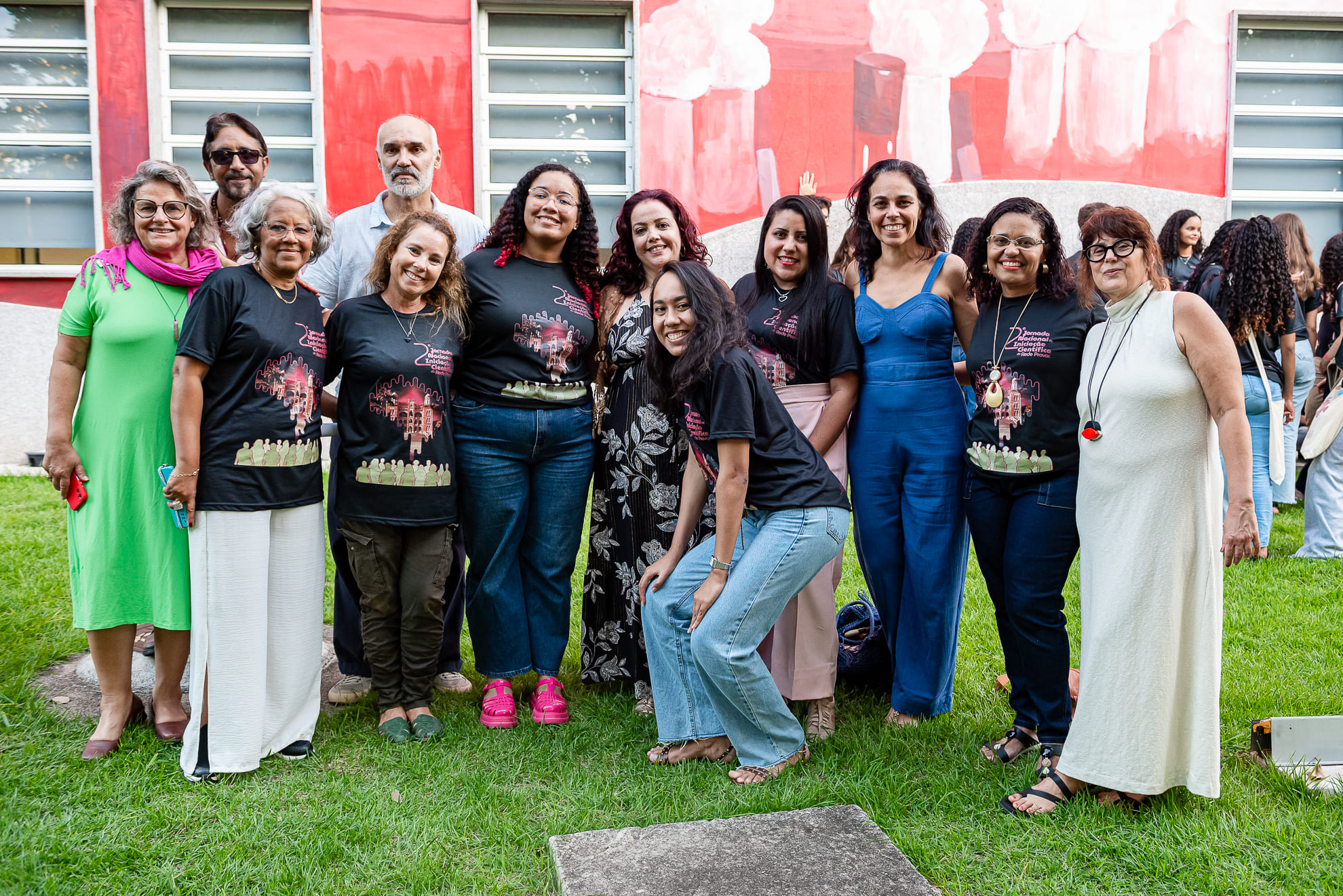Foto em grupo da equipe de profissionais do Provoc, responsáveis pela organização da 2ª Jornada Nacional de Iniciação Científica da Rede Provoc na Fiocruz. As 12 pessoas posam sorrindo em um gramado, em frente a uma parede com pintura artística em tons de vermelho. Algumas estão com a camiseta preta oficial do evento, com a arte da Jornada estampada. As demais usam roupas coloridas e variadas, como vestidos, macacões e calças. O clima é de confraternização e alegria.