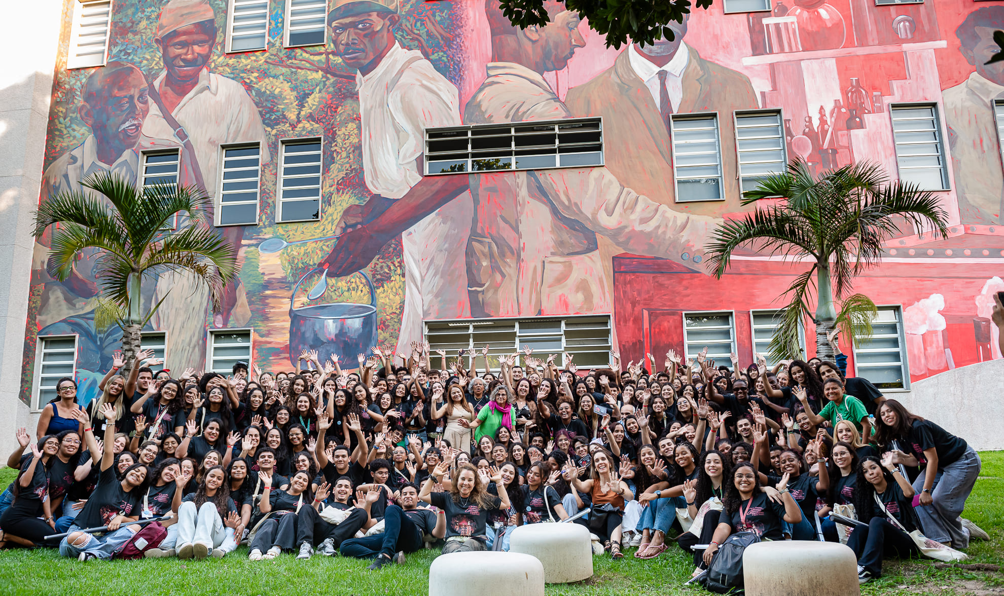 Grande grupo de estudantes do ensino médio, de diferentes estados do Brasil, posa sorrindo e com os braços erguidos em frente ao prédio da Escola Politécnica de Saúde Joaquim Venâncio, da Fiocruz. Ao fundo, há um mural com pintura de trabalhadores e elementos relacionados à saúde e à ciência.