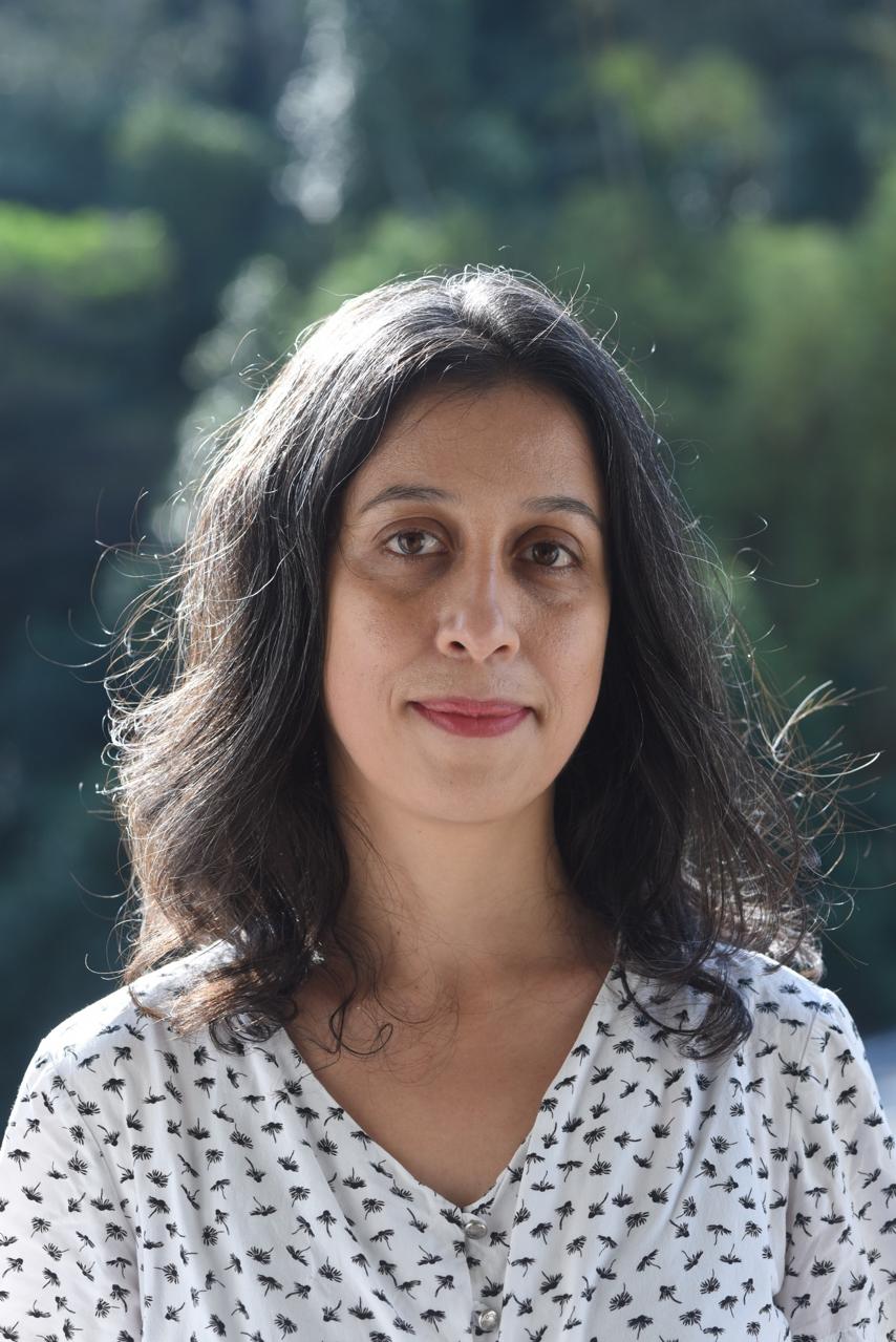 Retrato de uma pessoa de frente, com cabelo na altura dos ombros, usando uma blusa clara com pequenas estampas. O fundo é verde e desfocado, indicando um ambiente ao ar livre.
